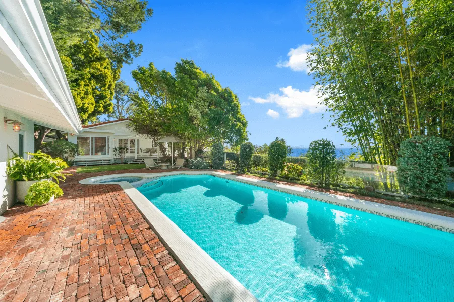 Back patio and pool area of a light gray craftsman style home in Malibu with lush green grass, small rectangular pool, large windows, palm trees, and neighboring house.