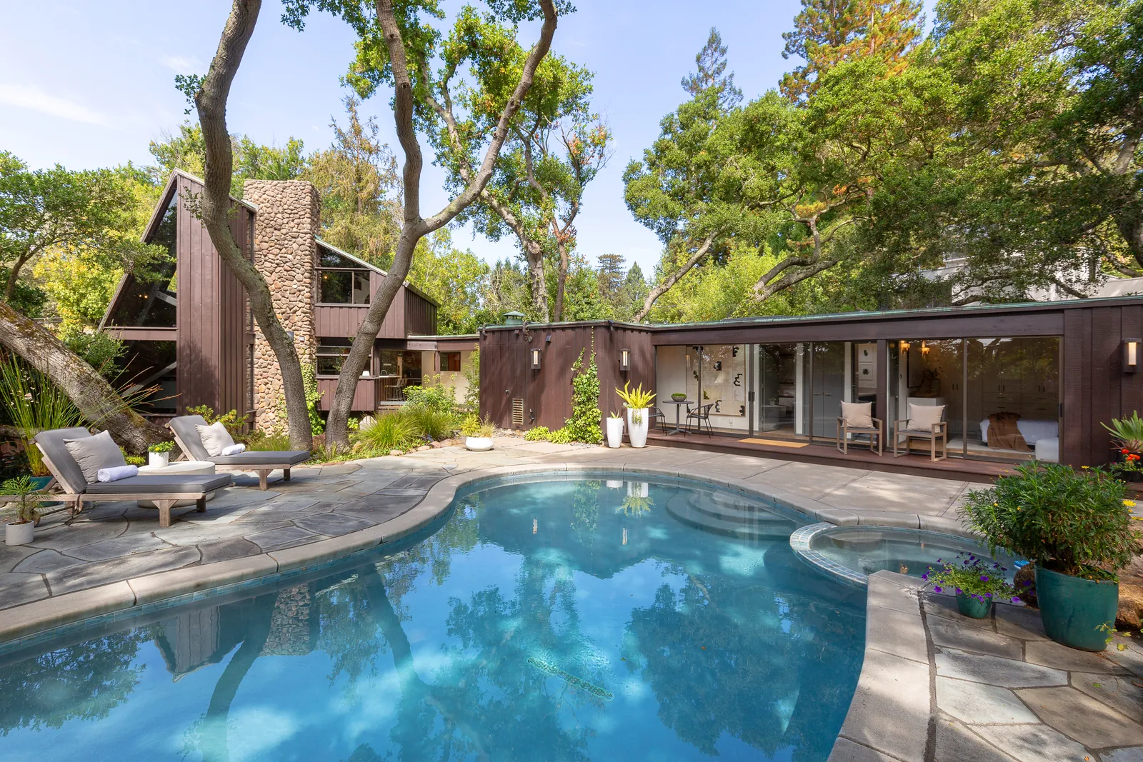 Mid-century modern East Bay home with dark wood siding and a stone chimney, set among mature oak trees behind a backyard swimming pool