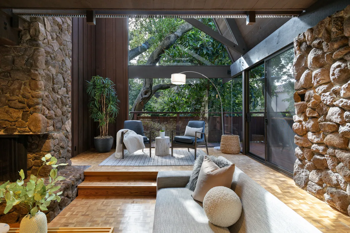 Living room with floor-to-ceiling glass walls and stone accents opening to tree canopy