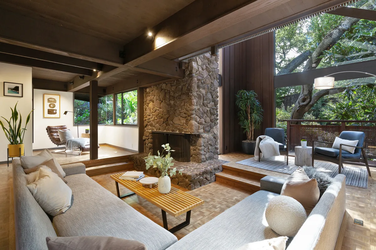 Living room in a mid-century East Bay home with a stone fireplace and oak trees outside