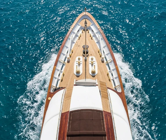 Aerial view looking straight down the bow of a polished wooden luxury yacht moving through deep blue open ocean water, with white wake trailing behind