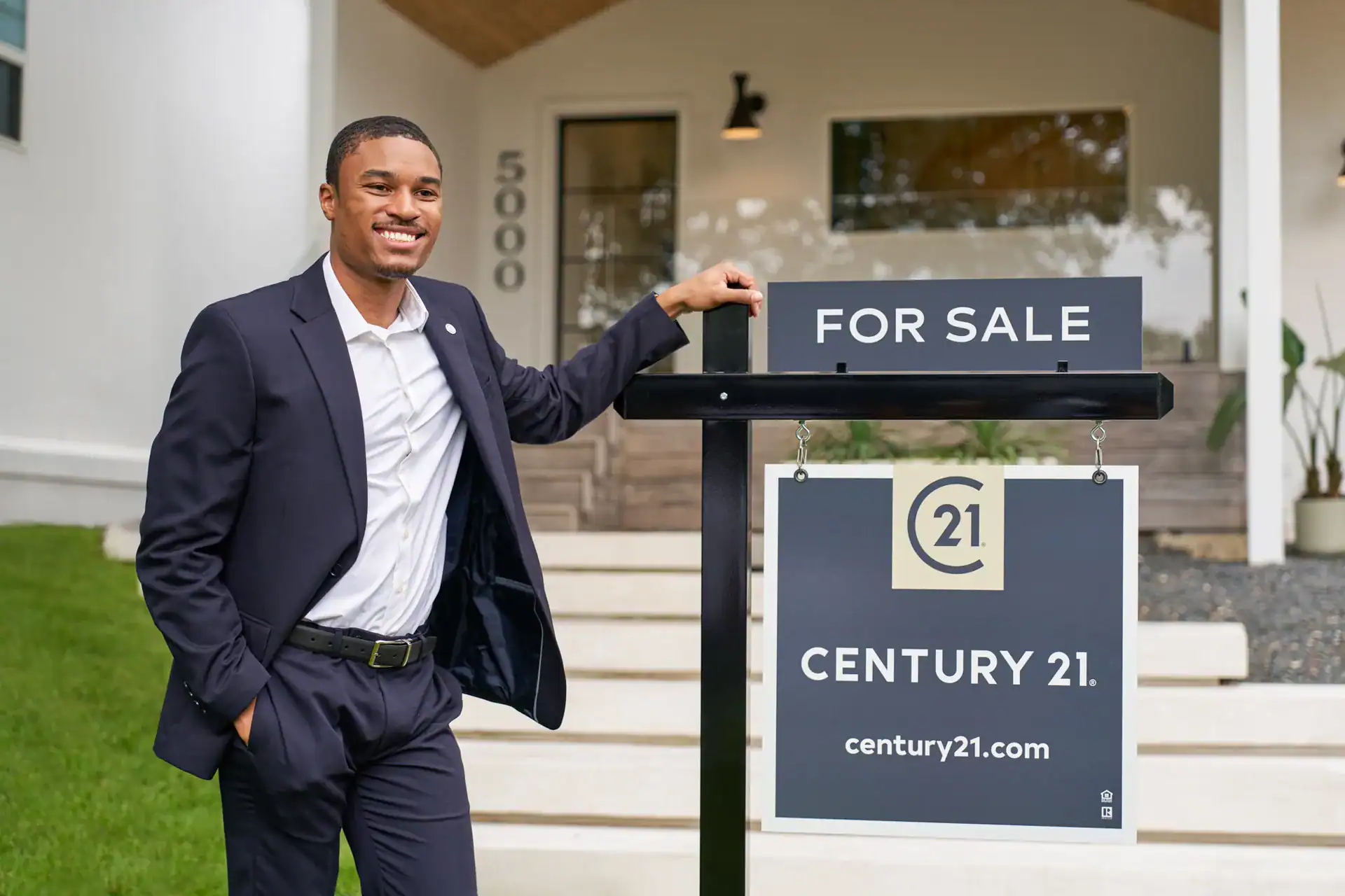 Smiling Century 21 agent leaning on a FOR SALE sign