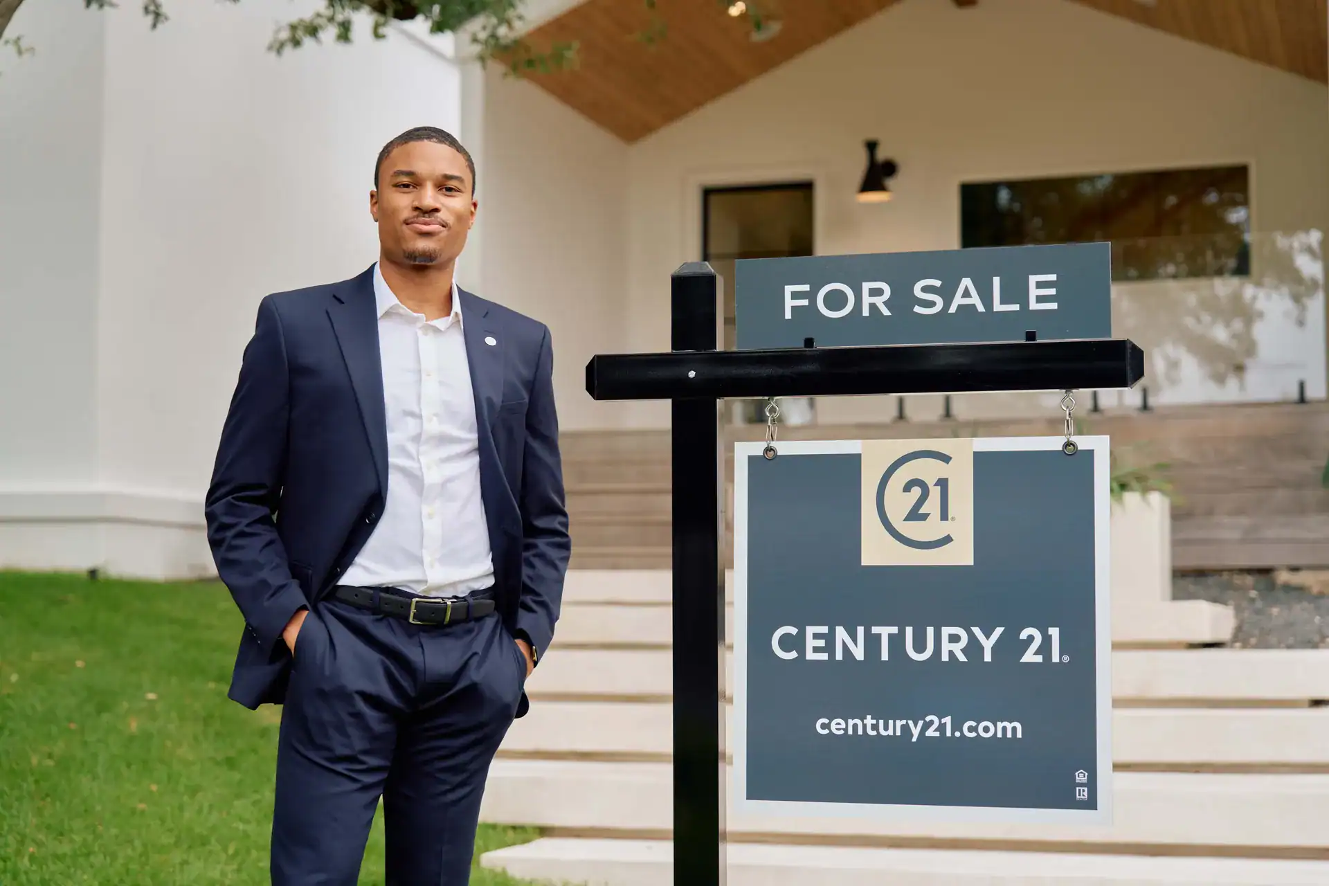 Century 21 agent in a navy suit beside a FOR SALE sign