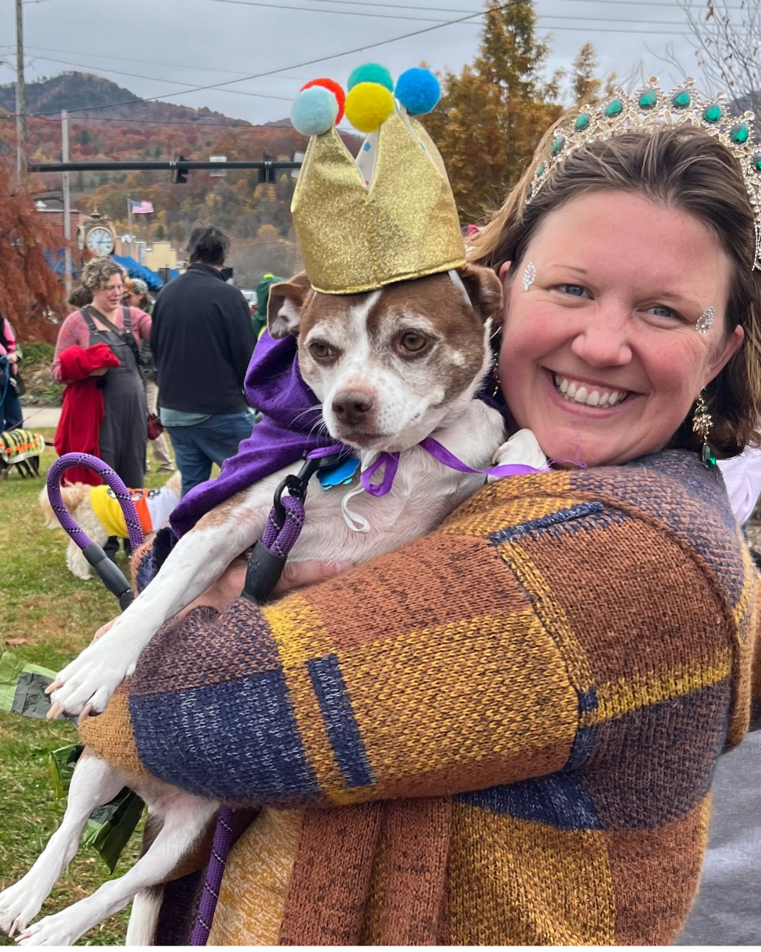 Woman with with sparkled face holds brown and white chihuahua with a purple cape and golden crown with Black Mountain Town Square, Clock and community members with their dressed up pets in the background, all against the mountain ranges of Black Mountain, North Carolina