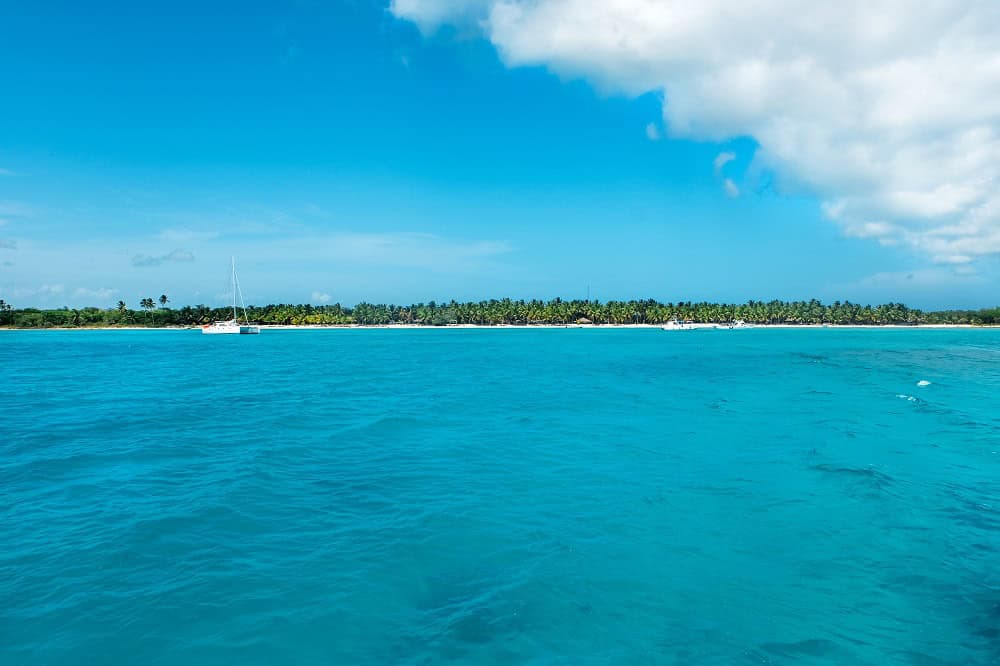 Bright blue ocean water with a distant shoreline lined with palm trees under a partly cloudy sky; a few boats are visible near the shore.