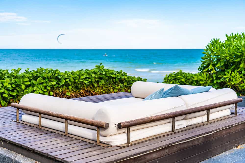 A modern white outdoor lounge sofa with blue pillows sits on a wooden deck in Albany, Bahamas, overlooking the ocean, with lush greenery and a clear blue sky in the background—perfect for luxurious real estate living.