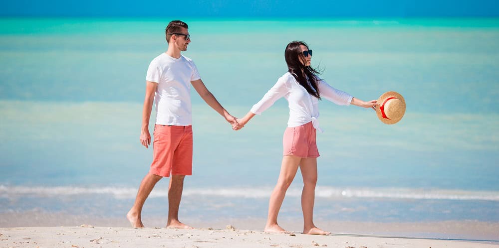 A man and woman in matching white shirts and coral shorts hold hands while walking along a sandy beach with turquoise water in the background.