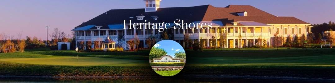 A wide-angle, panoramic image showing the large, multi-story Heritage Shores community clubhouse with white balconies at sunset. In the foreground is a golf course green beside a body of water, and a small circular inset graphic shows the Heritage Shores community entrance sign.