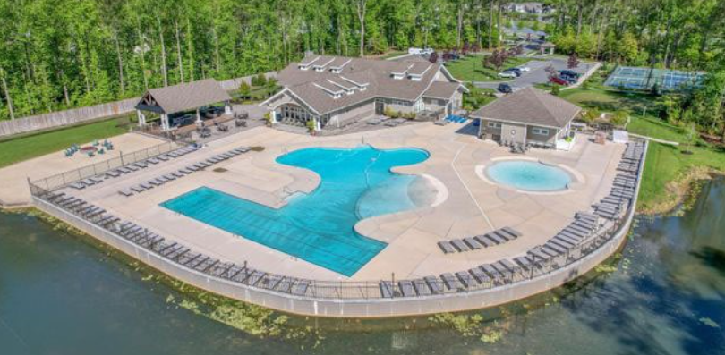 An aerial view of the amenity center at The Estuary in Frankford, DE, situated beside a lake and surrounded by trees. The complex features a large, custom-shaped swimming pool, a smaller circular wading pool, and an expansive concrete deck lined with lounge chairs. In the background, a multi-gabled clubhouse, a covered outdoor pavilion, and tennis courts complete the recreational facilities.