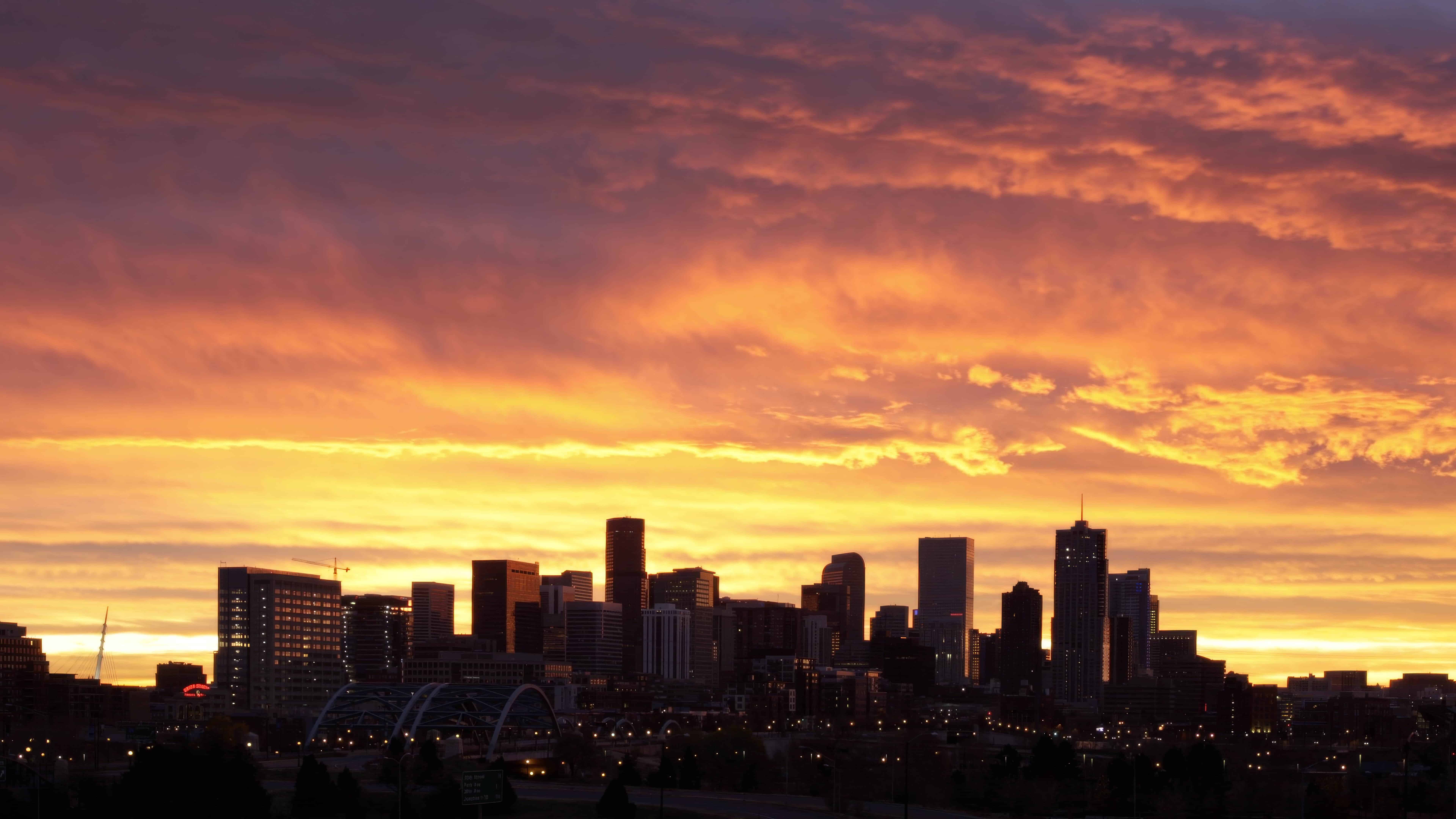 Downtown Denver skyline silhouetted against an orange and pink sunset from a pedestrian bridge