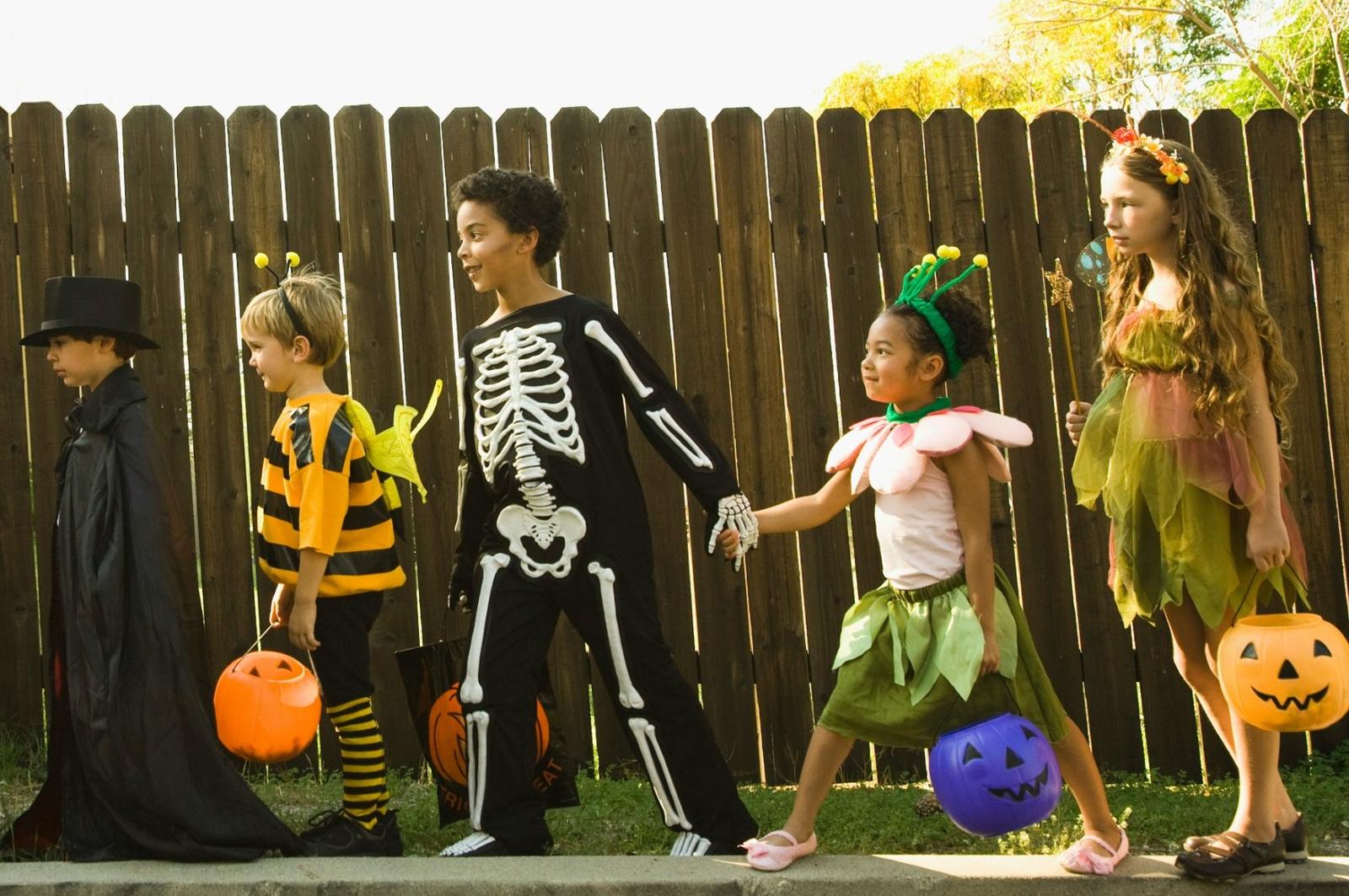 Children in Halloween costumes, including a skeleton, a bee, a flower, and a fairy, walking with pumpkin buckets
