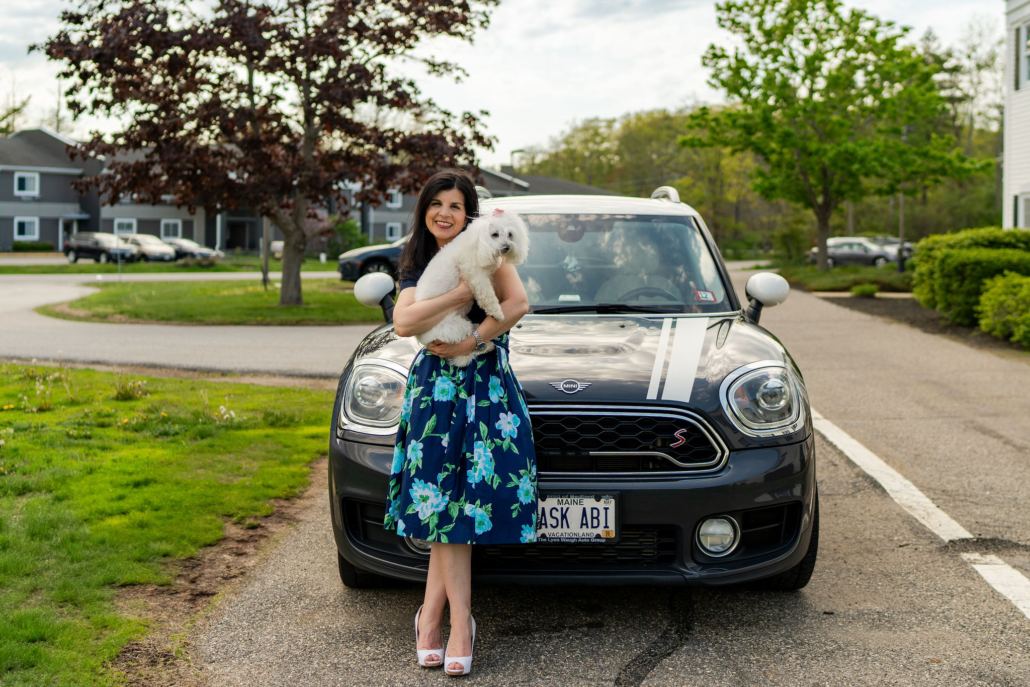 Abigail Douris holding a small white dog in front of her car with an ASK ABI license plate