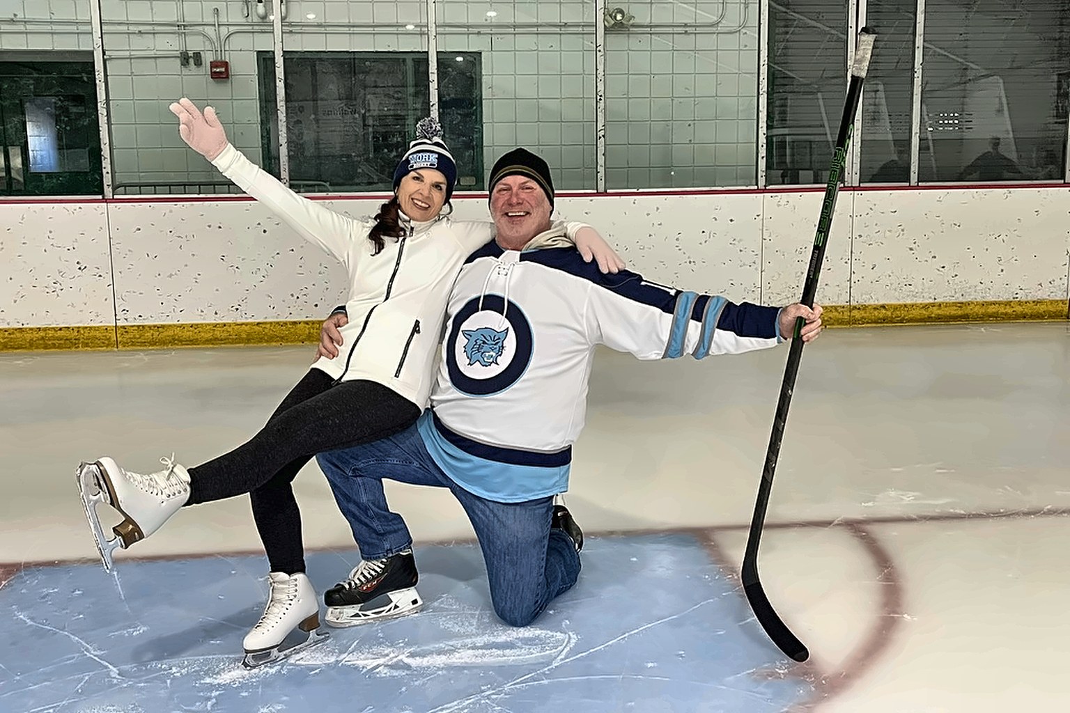 Peter Douris kneeling on the ice with a hockey stick while Abigail Douris, in figure skates, sits on his knee